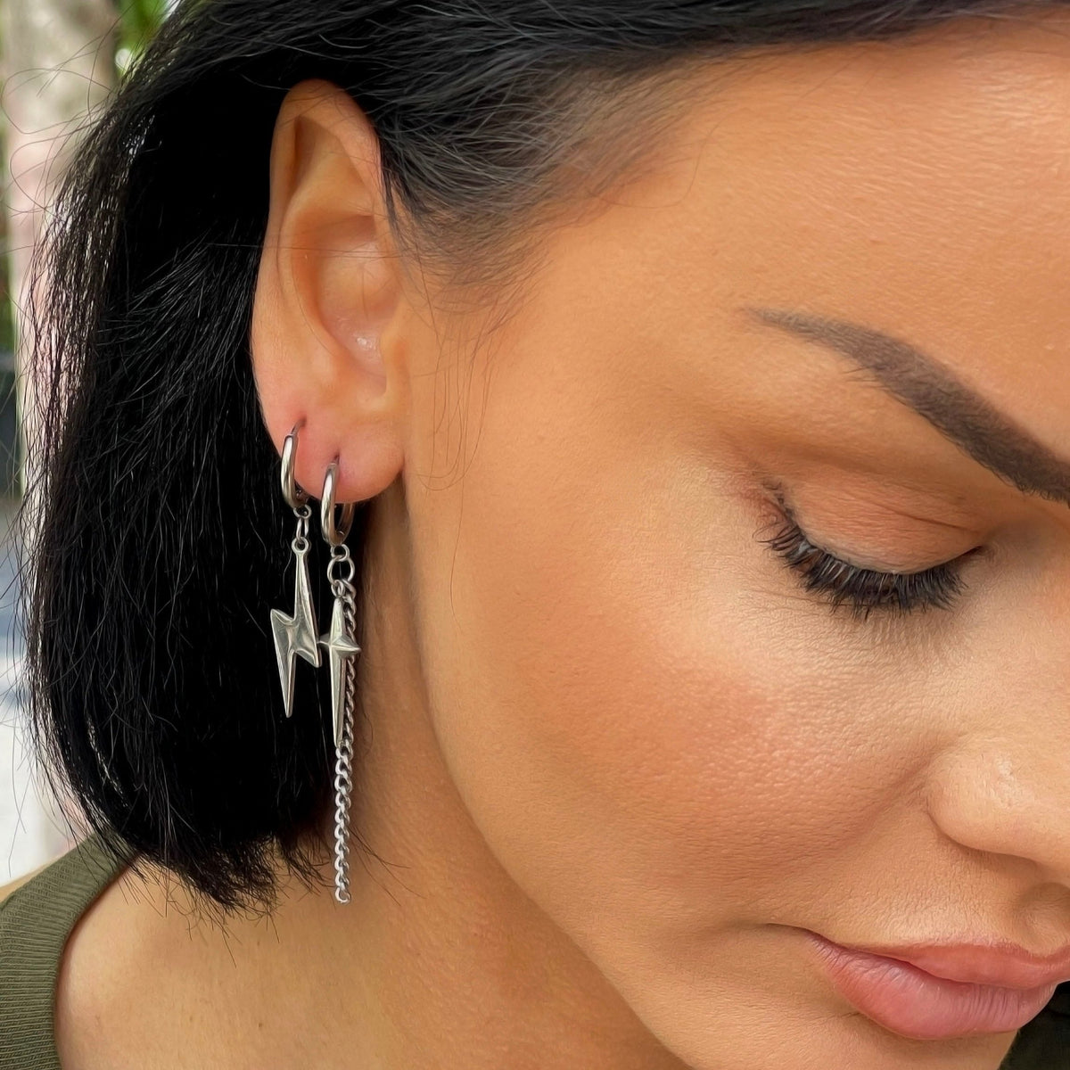Close-up of a woman wearing Atlas Lightning Bolt mismatch dangle earrings featuring a cross and lightning bolt design in stainless steel